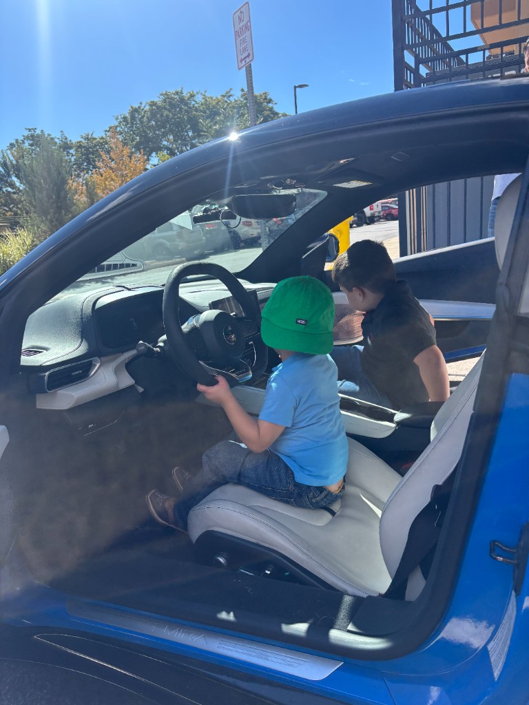 Child in a green hat holding the wheel of a blue Lotus, with another child beside them