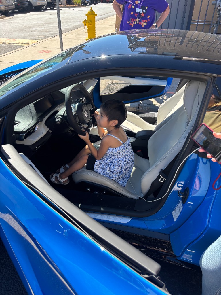 Child in a floral dress sitting at the wheel of a blue sports car